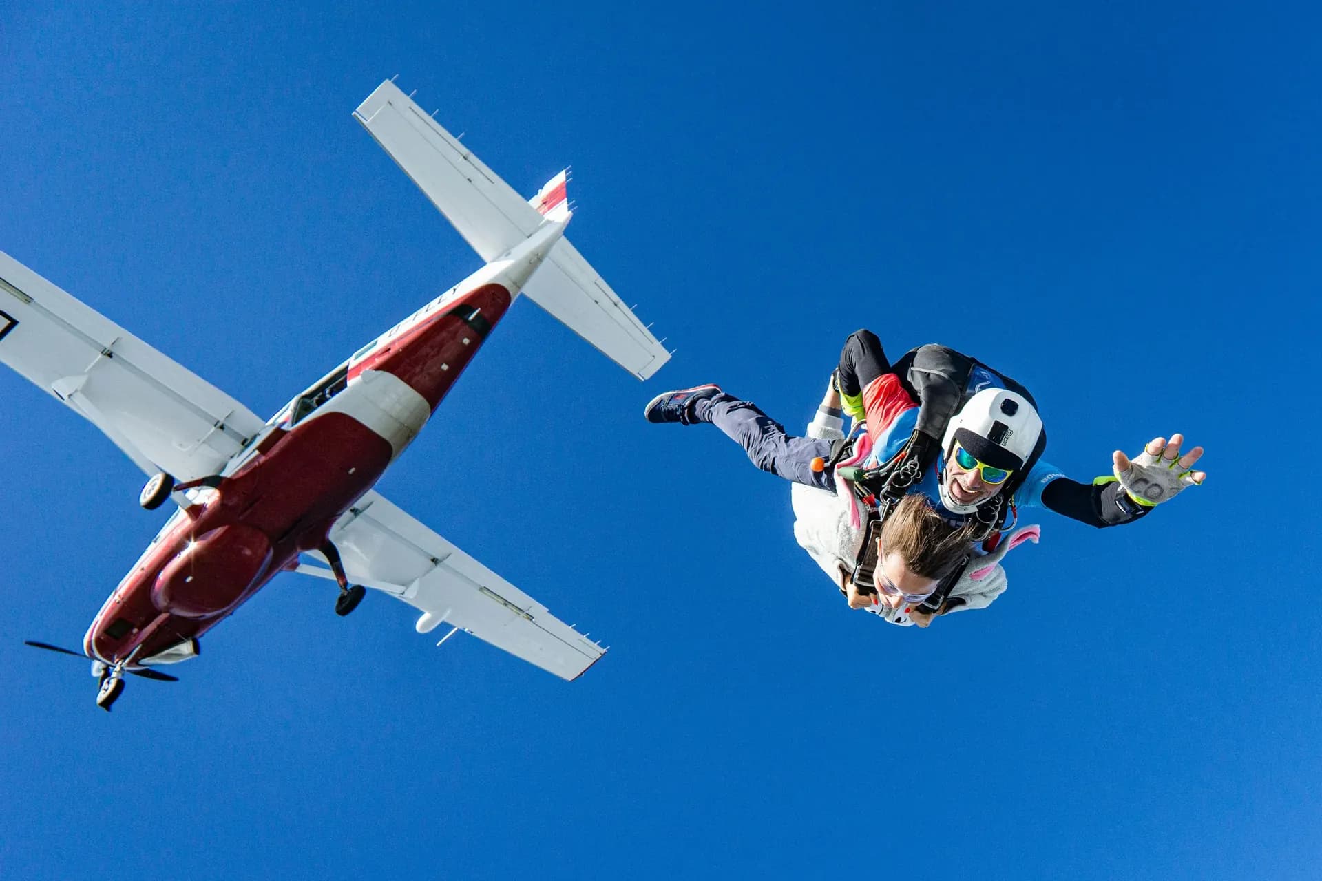 Tandem skydivers in freefall with the aircraft above, bright blue sky over Asia